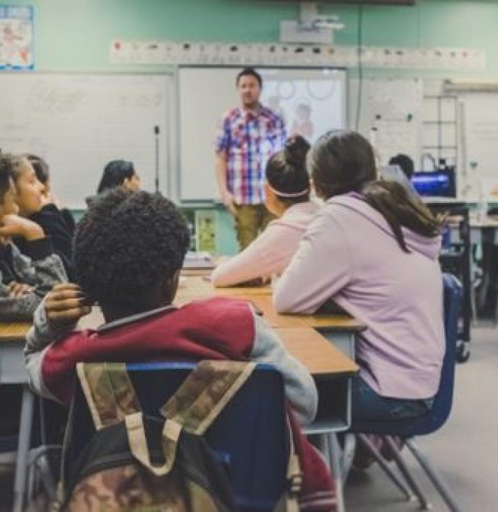 A man stood at the front of a classroom while seated students listen attentively