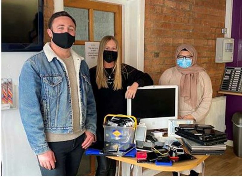 3 people stood around a table filled with used computer electricals that are to be recycled for continued use not thrown away.